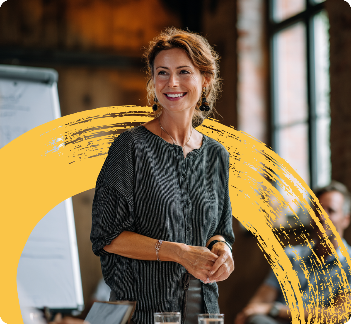 Smiling woman standing and speaking in a meeting room with a flip chart and seated people in the background.