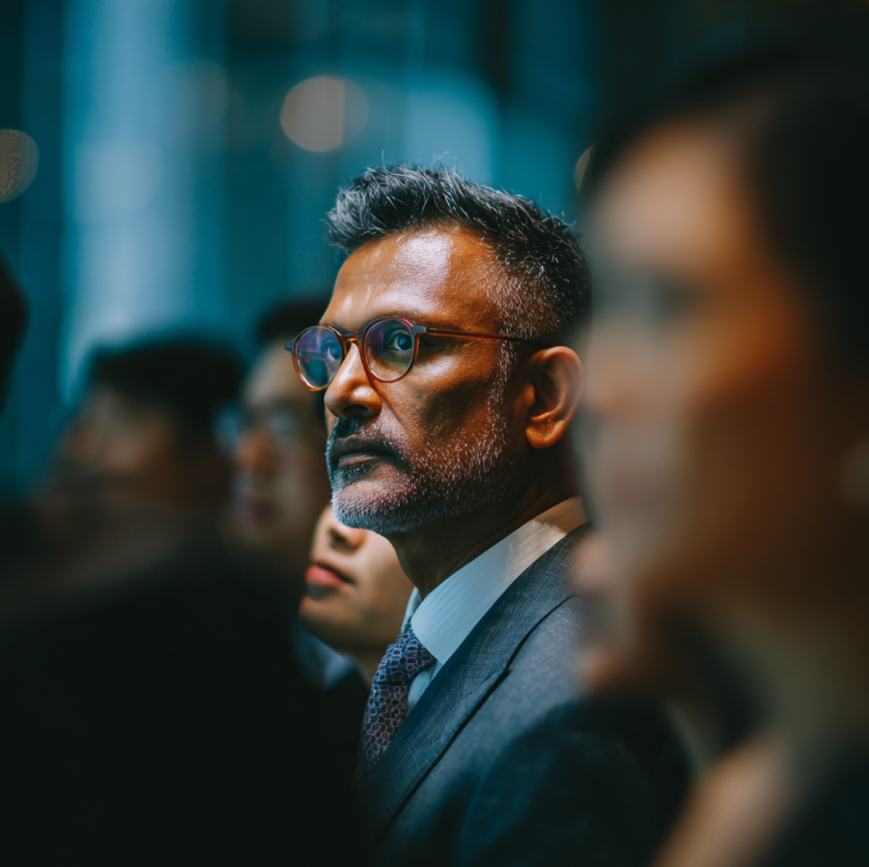 Middle-aged man with glasses and a beard, dressed in a suit, attentively looking forward in a dimly lit setting with blurred people in the background.