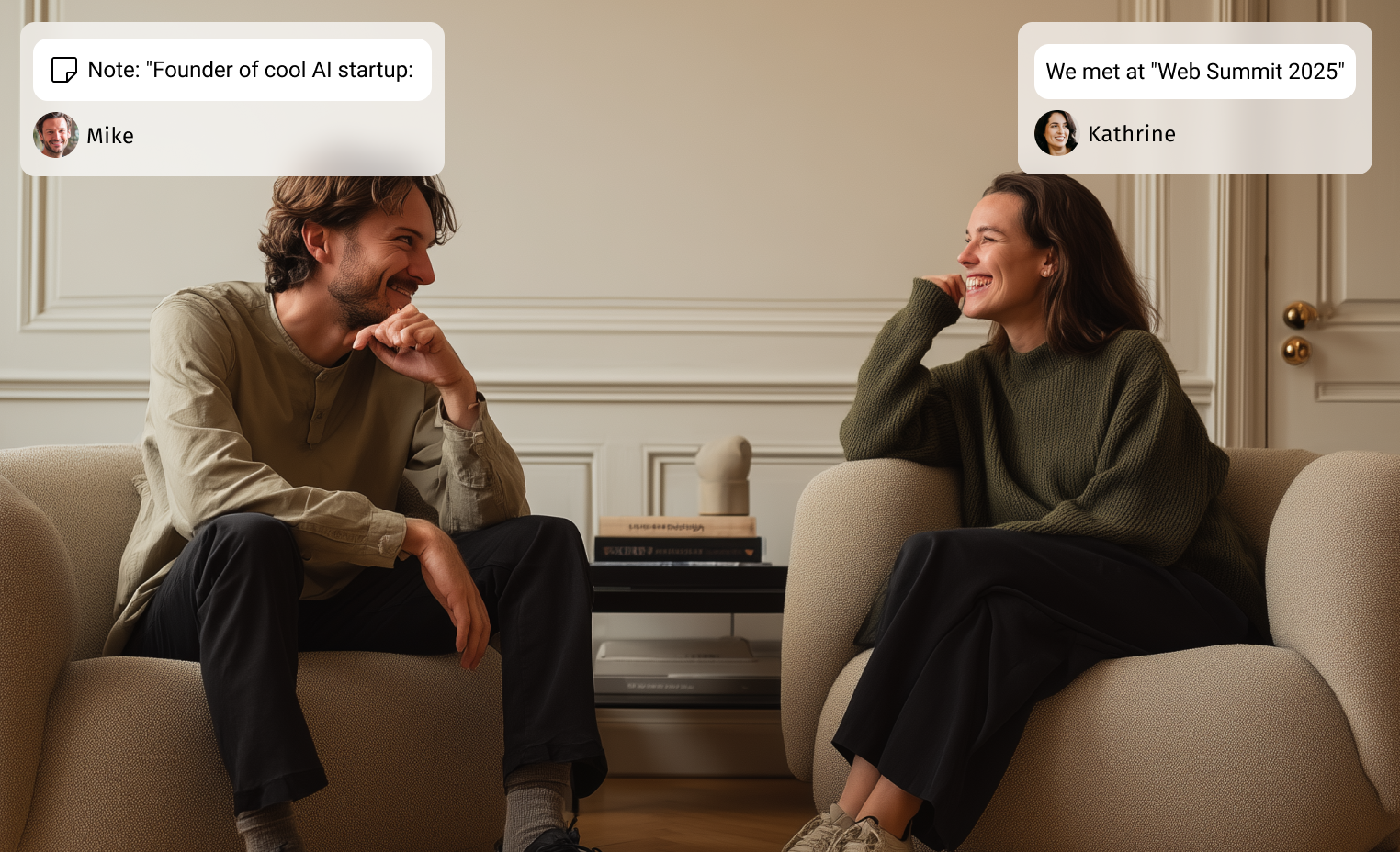 A man and a woman sitting in beige armchairs facing each other, smiling and engaged in conversation in a room with beige walls and a small table with books in the background.