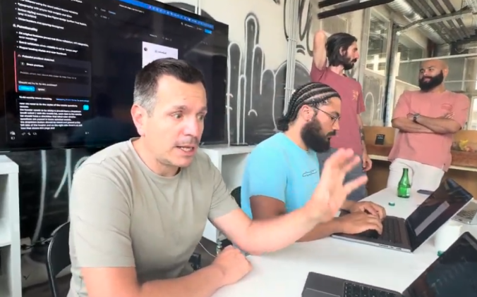 Four men collaborating in an office setting, with two seated typing on laptops and two standing in conversation, a screen displaying text in the background.